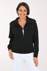A smiling woman wearing a black zip-up jacket over a white top and white pants, standing against a plain light background.