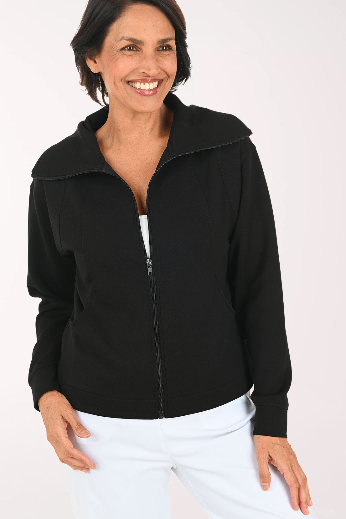 A smiling woman wearing a black zip-up jacket over a white top and white pants, standing against a plain light background.