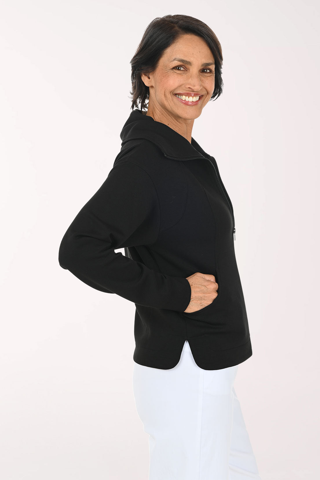 A smiling woman with dark hair is shown from the side/back wearing a black long-sleeve hooded top and white pants against a plain light background.
