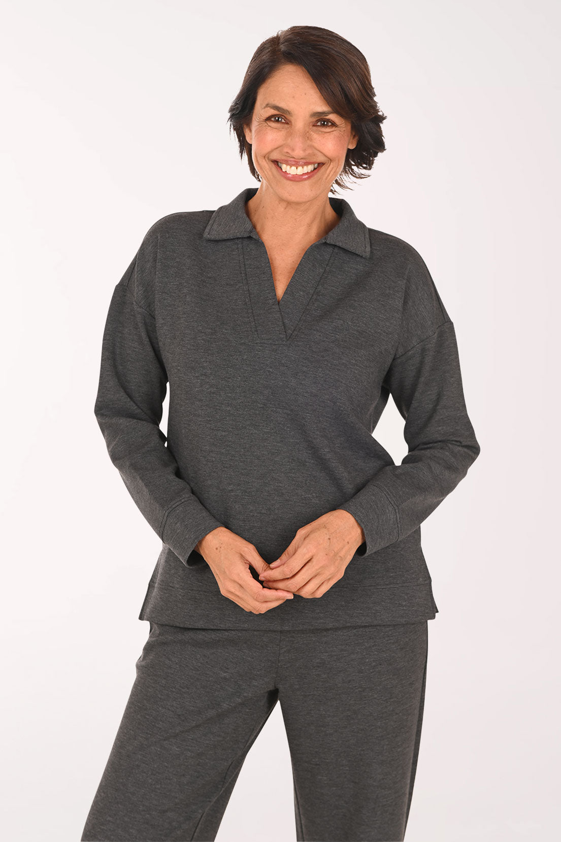 A smiling woman wearing a dark gray long-sleeve polo-style top and matching dark gray pants, standing against a plain light background.