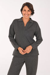 A smiling woman wearing a dark gray long-sleeve polo-style top and matching dark gray pants, standing against a plain light background.