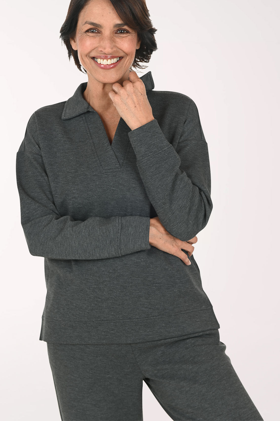 A smiling woman wearing a dark gray long-sleeve polo-style top and matching dark gray pants, standing against a plain light background.