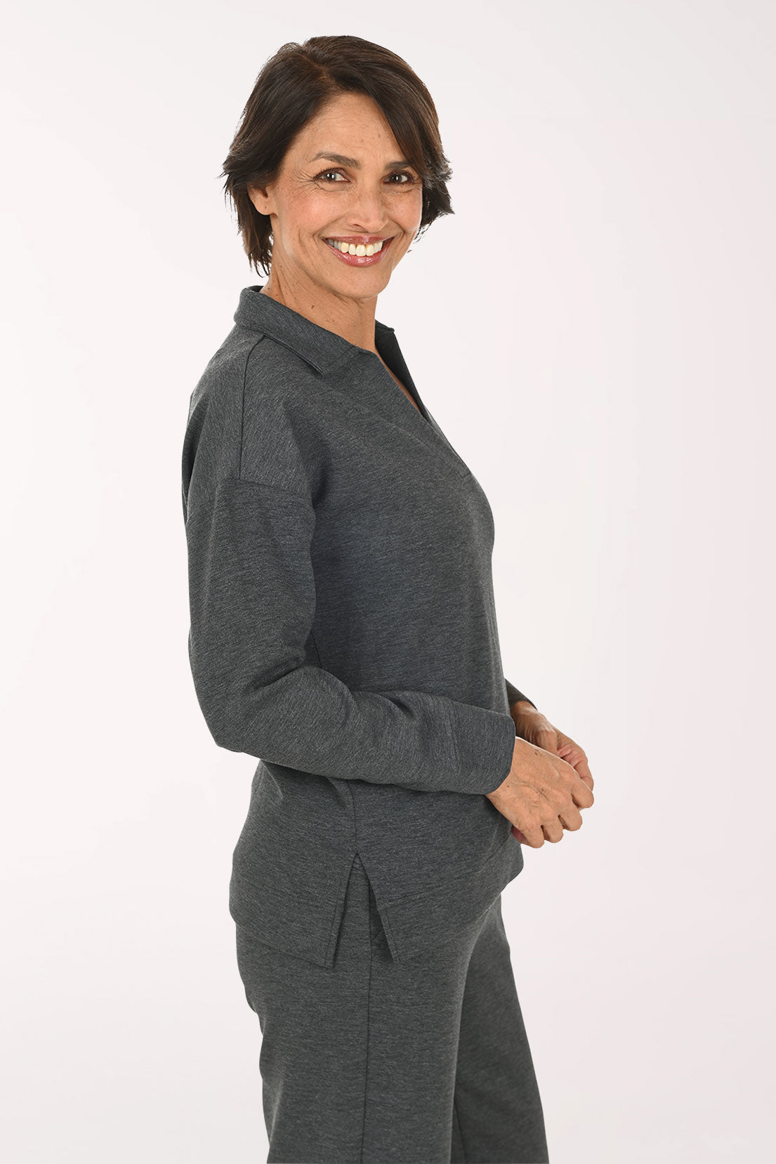 A smiling woman wearing a dark gray long-sleeve polo-style top and matching dark gray pants, standing against a plain light background.