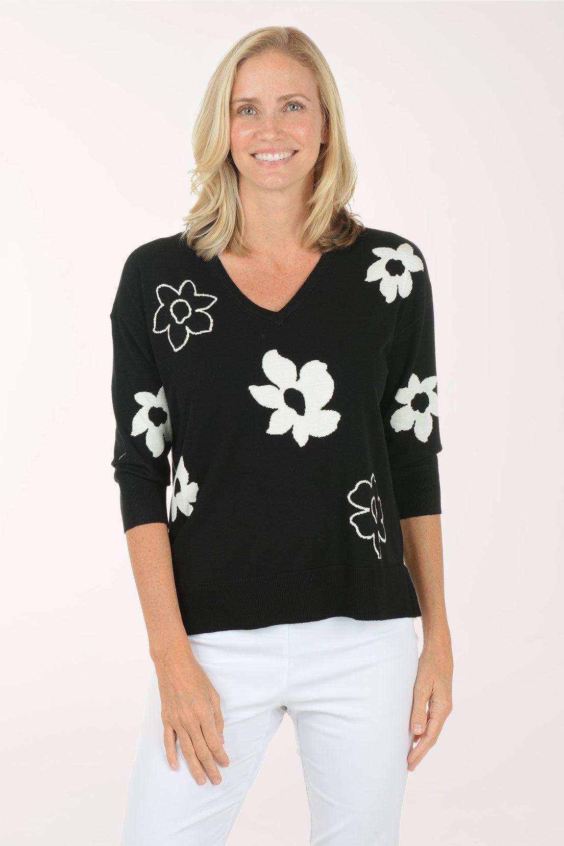A smiling blonde woman wearing a black V-neck knit sweater with large white floral patterns and white pants, standing against a plain light background.
