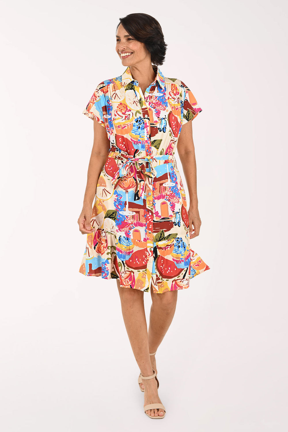 A woman modeling a short-sleeve collared shirt dress with a colorful abstract print, featuring a tie waist and ruffled hem, set against a plain white studio background.