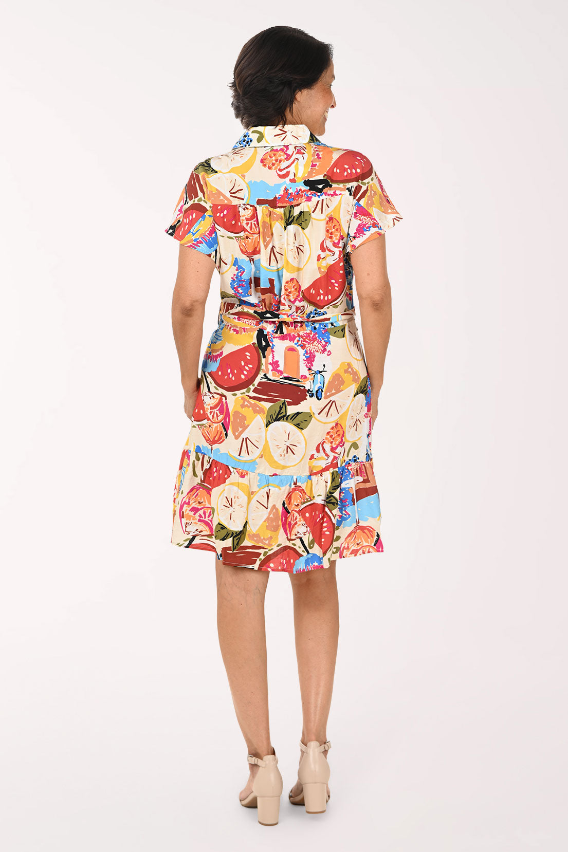 A woman modeling a short-sleeve collared shirt dress with a colorful abstract print, featuring a tie waist and ruffled hem, set against a plain white studio background.