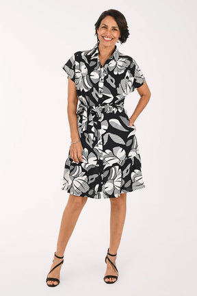 A woman modeling a short-sleeve black and white tropical floral print dress with a tie waist, paired with black strappy high-heeled sandals, against a plain white studio background.