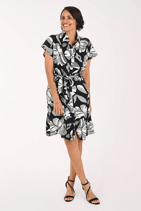 A woman modeling a short-sleeve black and white tropical floral print dress with a tie waist, paired with black strappy high-heeled sandals, against a plain white studio background.