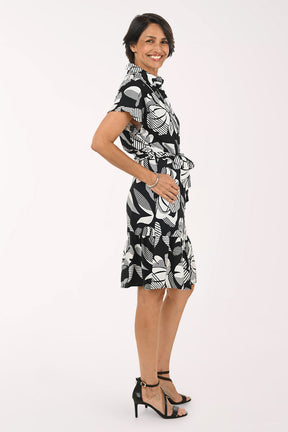A woman modeling a short-sleeve black and white tropical floral print dress with a tie waist, paired with black strappy high-heeled sandals, against a plain white studio background.