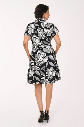 A woman modeling a short-sleeve black and white tropical floral print dress with a tie waist, paired with black strappy high-heeled sandals, against a plain white studio background.