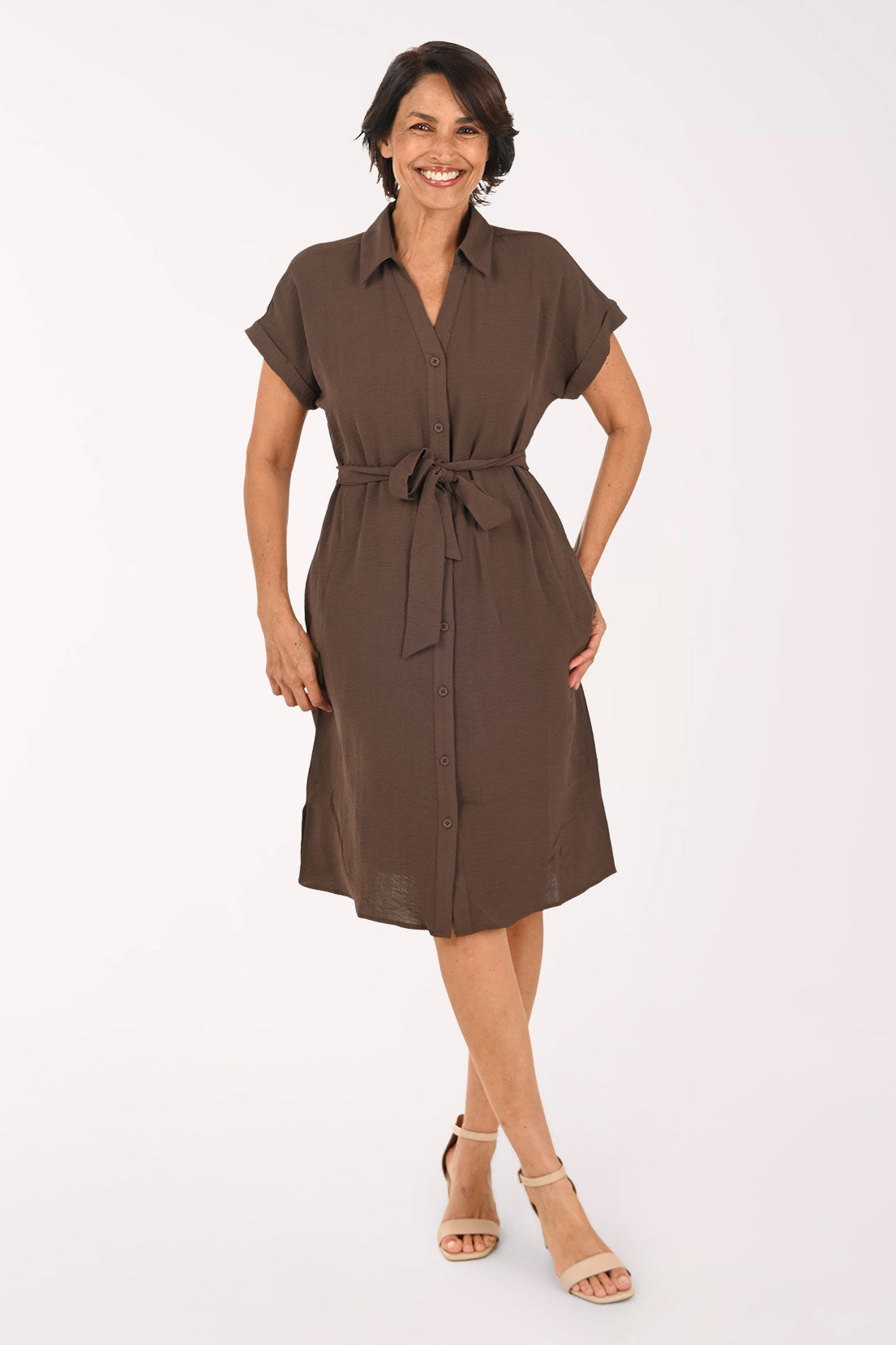 A woman modeling a brown short-sleeve shirt dress with a tie waist, standing against a plain white studio background.