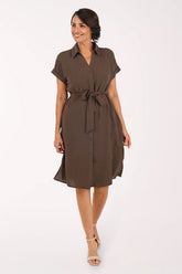 A woman modeling a brown short-sleeve shirt dress with a tie waist, standing against a plain white studio background.