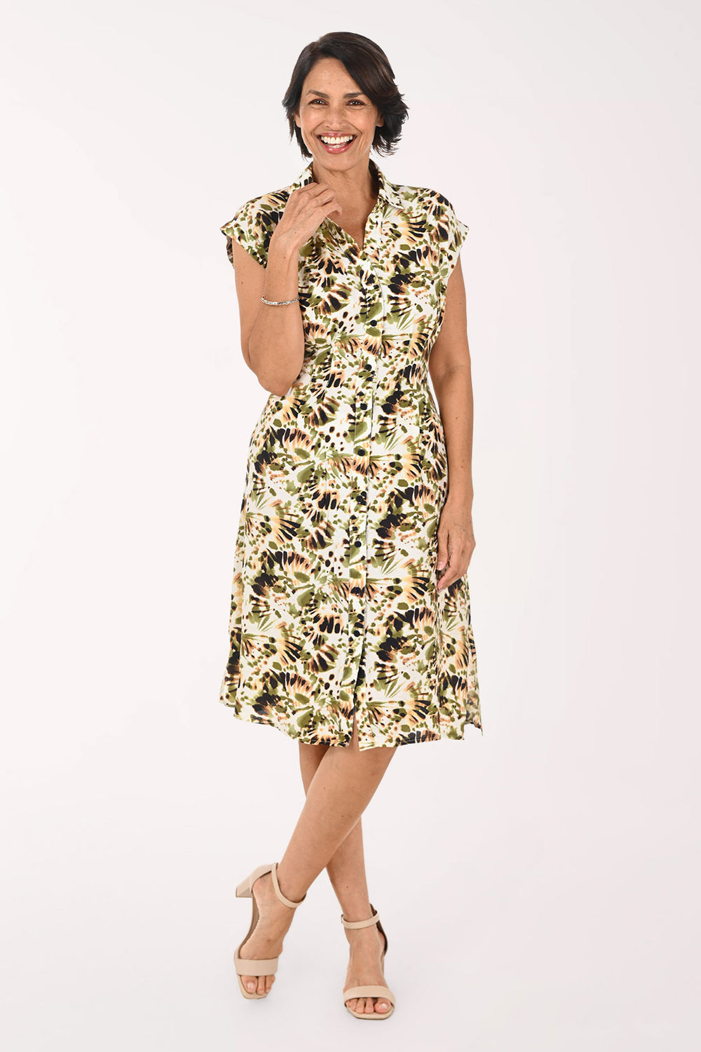A woman modeling a knee-length floral dress with a cream base and green, black, and brown botanical print, paired with beige heeled sandals against a plain white studio background.