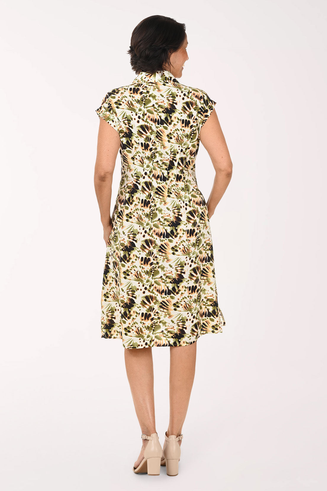 A woman modeling a knee-length floral dress with a cream base and green, black, and brown botanical print, paired with beige heeled sandals against a plain white studio background.
