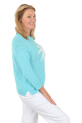 Woman wearing a light blue sweater with a palm tree design on a white background. Side shot.