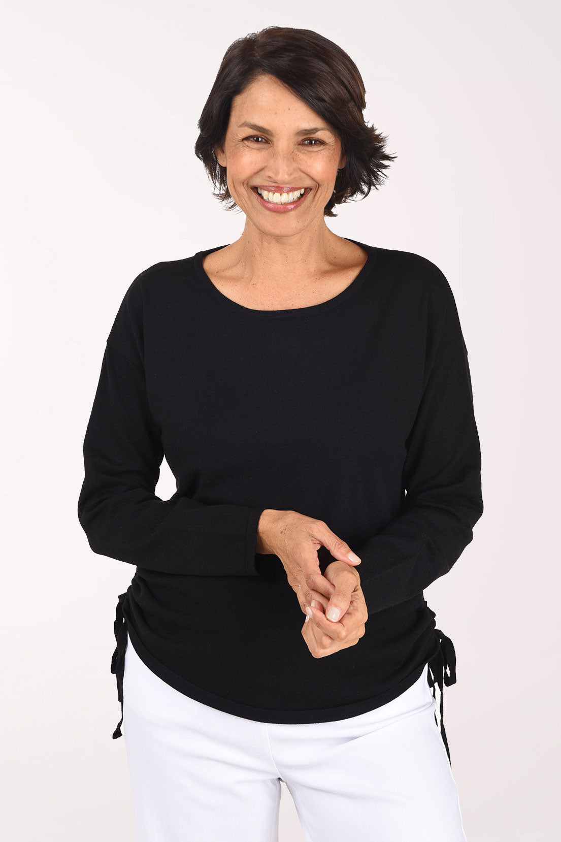 Smiling woman in a black long sleeve crew neck lightweight sweater with drawstring ruching gathered at both side seams, worn with white pants