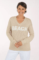 A woman wearing a beige V-neck knit sweater with the word 'BEACH' in large white letters across the chest, paired with white pants, against a plain light background.