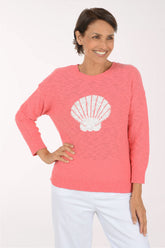 A smiling woman wearing a coral pink knit sweater with a large white seashell graphic on the front, paired with white pants, against a plain white studio background.