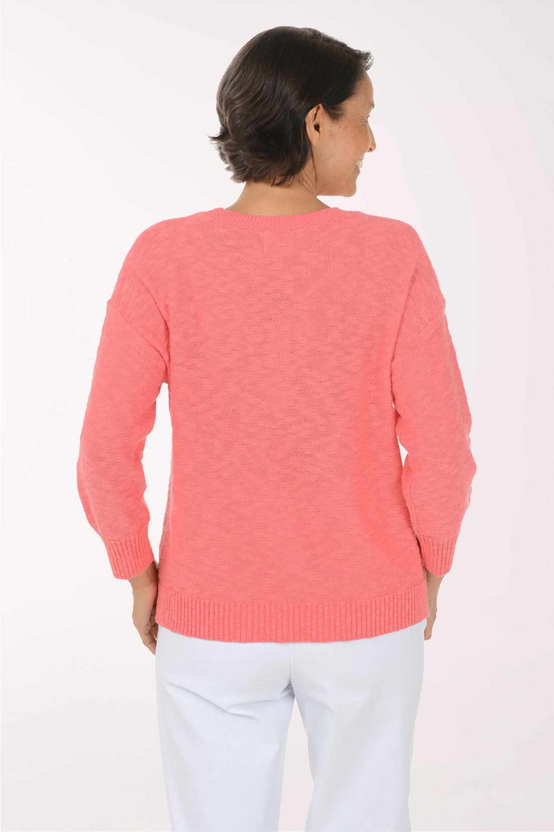 A smiling woman wearing a coral pink knit sweater with a large white seashell graphic on the front, paired with white pants, against a plain white studio background.
