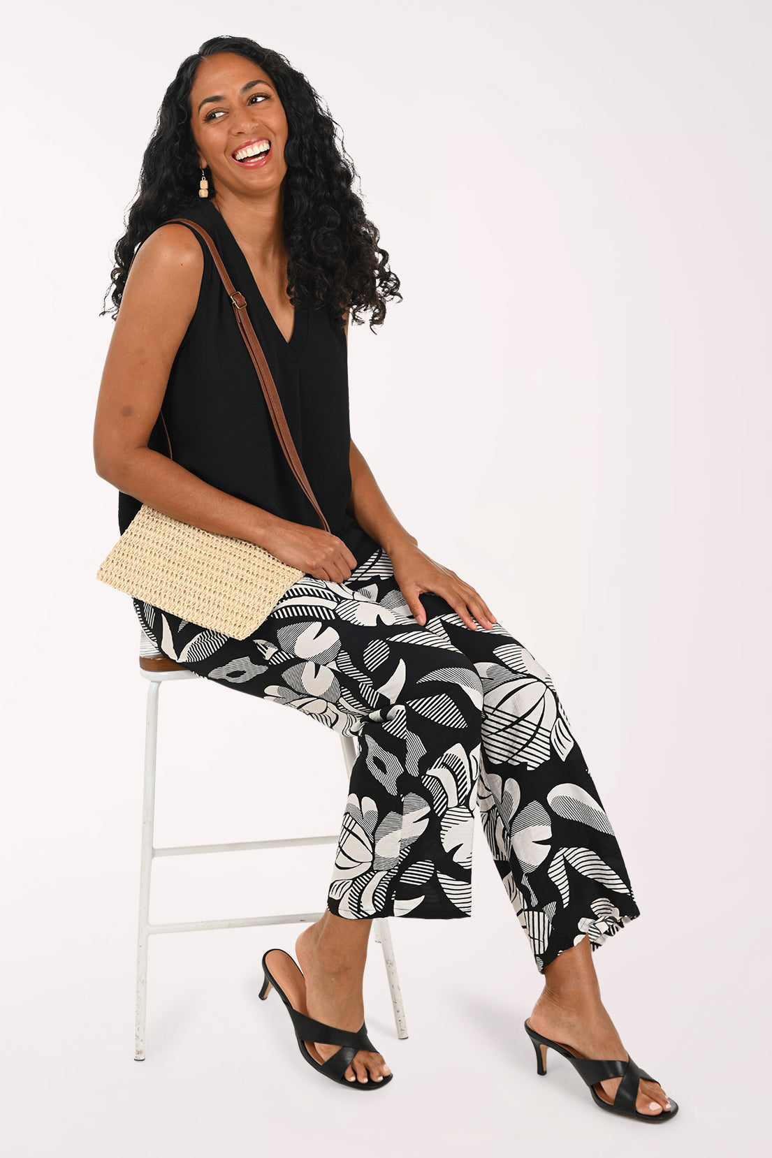 Woman sitting on a stool wearing a black sleeveless top and patterned pants, holding a beige handbag.