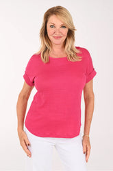 A smiling blonde woman wearing a bright pink short-sleeve crewneck top and white pants, standing against a plain white studio background.