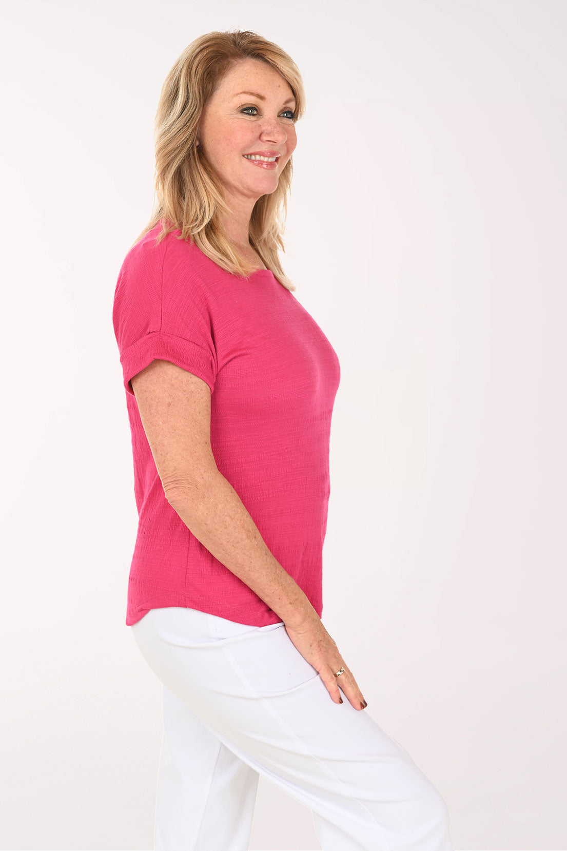 A smiling blonde woman wearing a bright pink short-sleeve crewneck top and white pants, standing against a plain white studio background - side shot