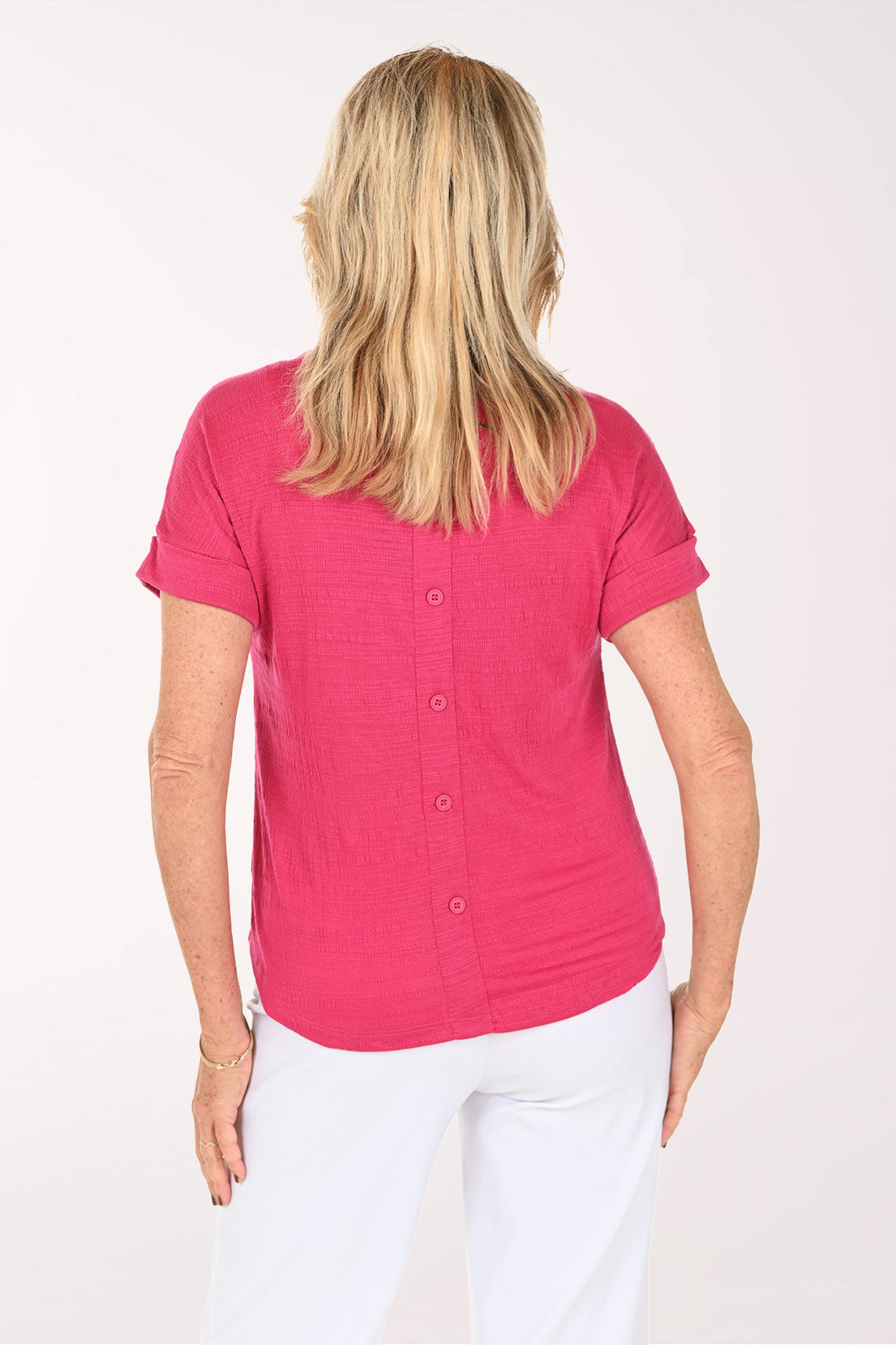 A smiling blonde woman wearing a bright pink short-sleeve crewneck top and white pants, standing against a plain white studio background - back shot