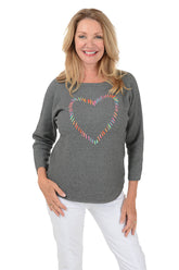 A smiling blonde woman wearing a heather gray long-sleeve top with a colorful heart outline graphic on the chest, paired with white pants, against a plain white background.