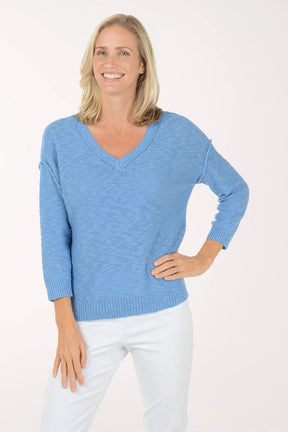 A woman wears a blue sweater and white pants against a light pink background