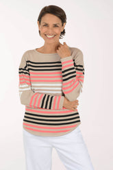 A smiling woman wearing a long-sleeve knit top with horizontal stripes in beige, black, and pink, paired with white pants, against a plain white studio background.
