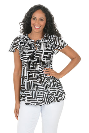 Woman wearing a black and white geometric patterned blouse with white pants on a white background.