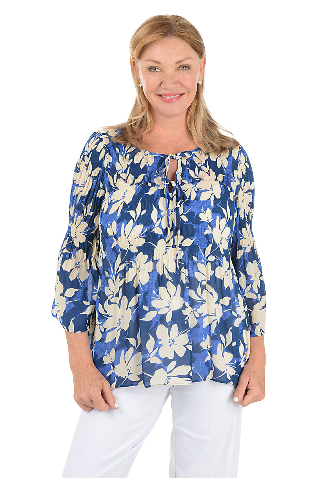 Woman wearing a blue floral blouse with white pants on a white background.