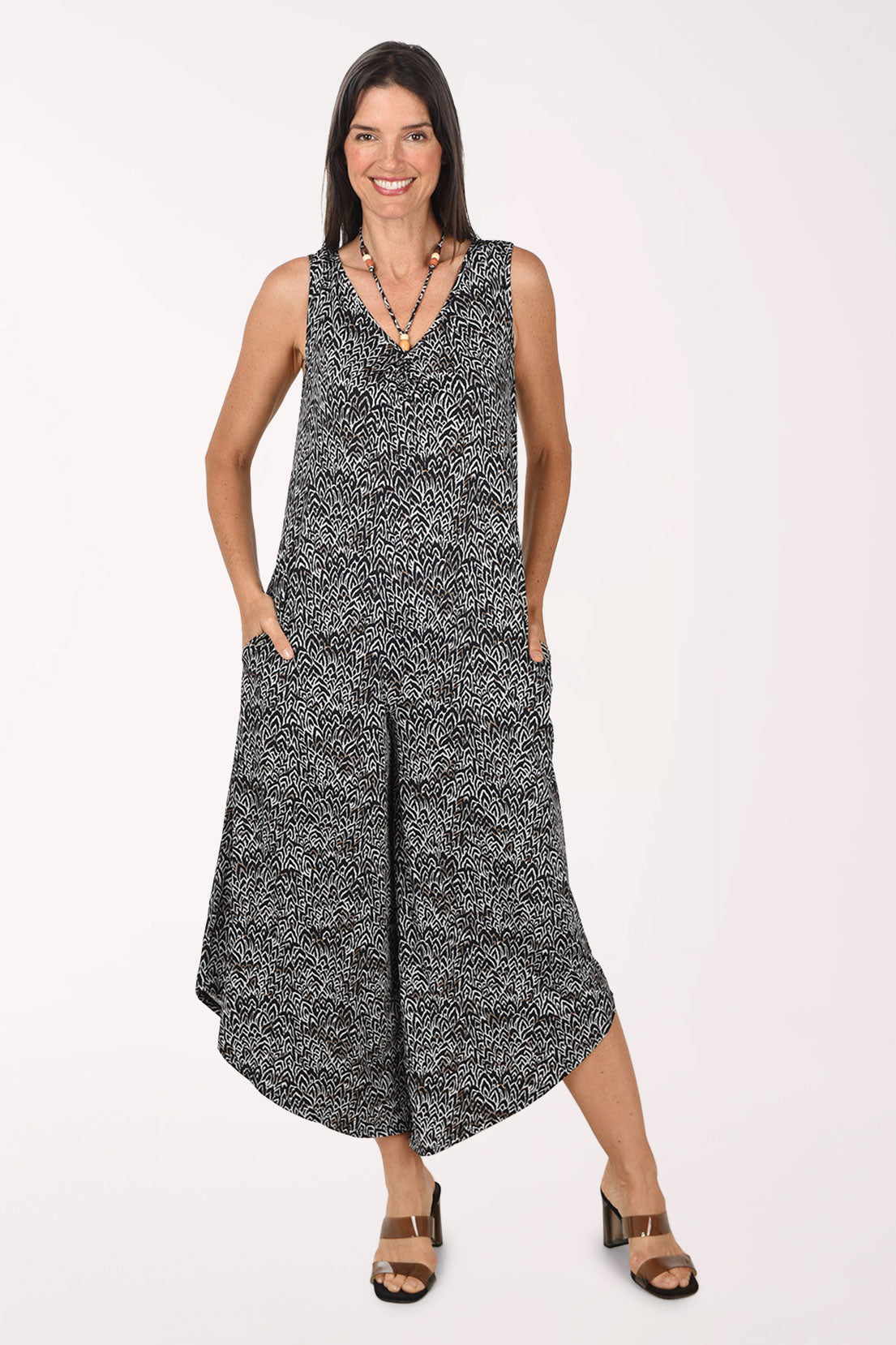 Front view of a model wearing the Sami and Jo Chevron Beaded Neck Sleeveless Jumpsuit in a black and white chevron print, with a V-neckline, hip pockets and wide cropped legs with an angled hem, worn with brown heeled sandals.