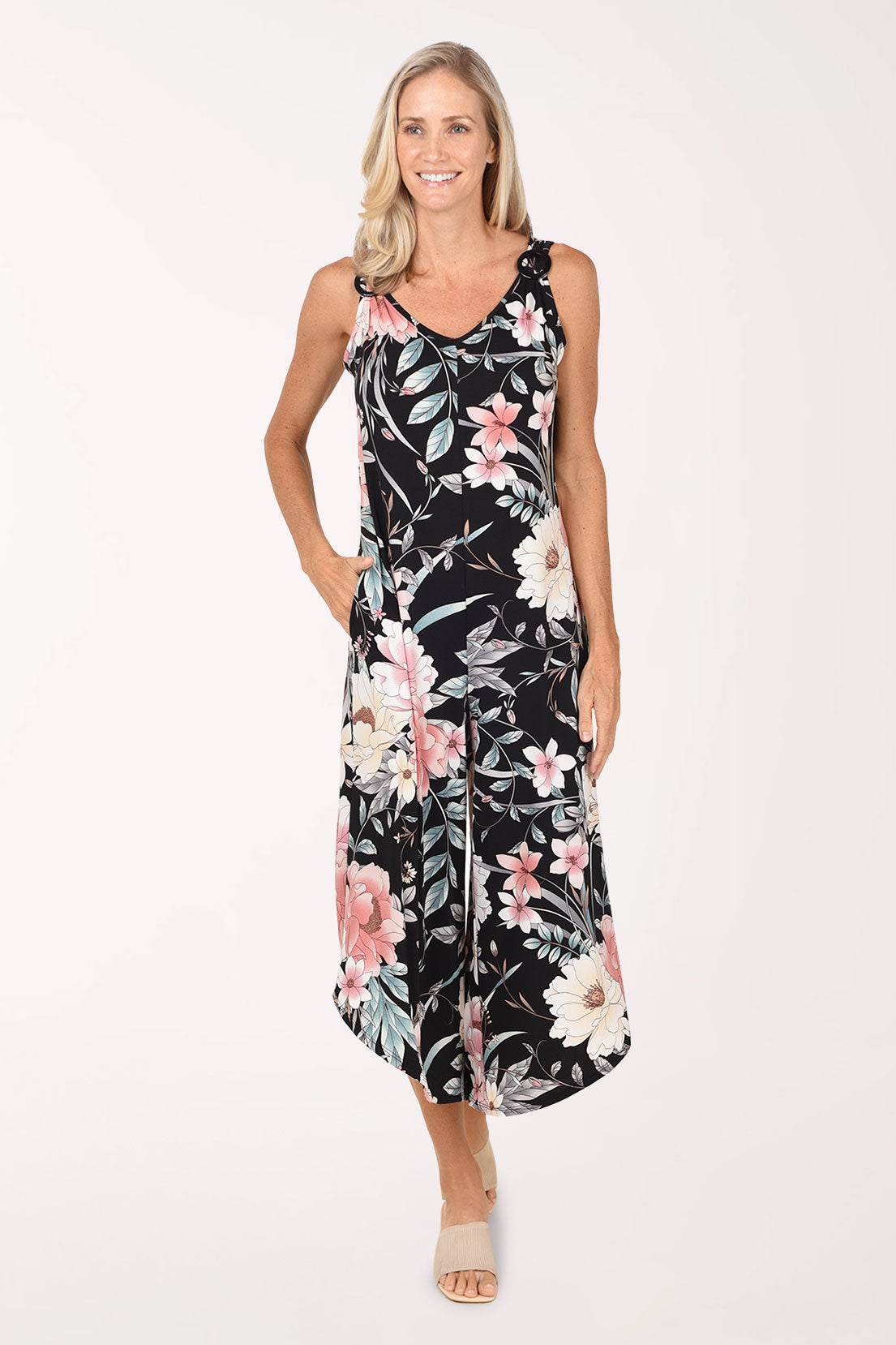 Front view of a model wearing the Sami and Jo Exotic Floral Ringed Strap Sleeveless Jumpsuit, a black jumpsuit with blush pink, cream and sage florals, a V-neckline and wide cropped legs with an angled hem, worn with nude sandals.