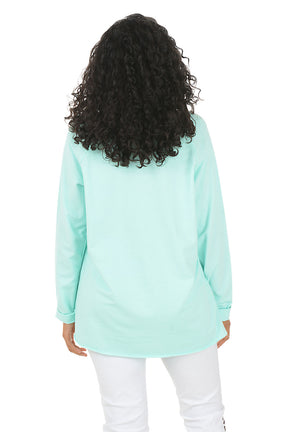 Woman wearing a light blue long-sleeve top and white pants on a white background. Back shot.
