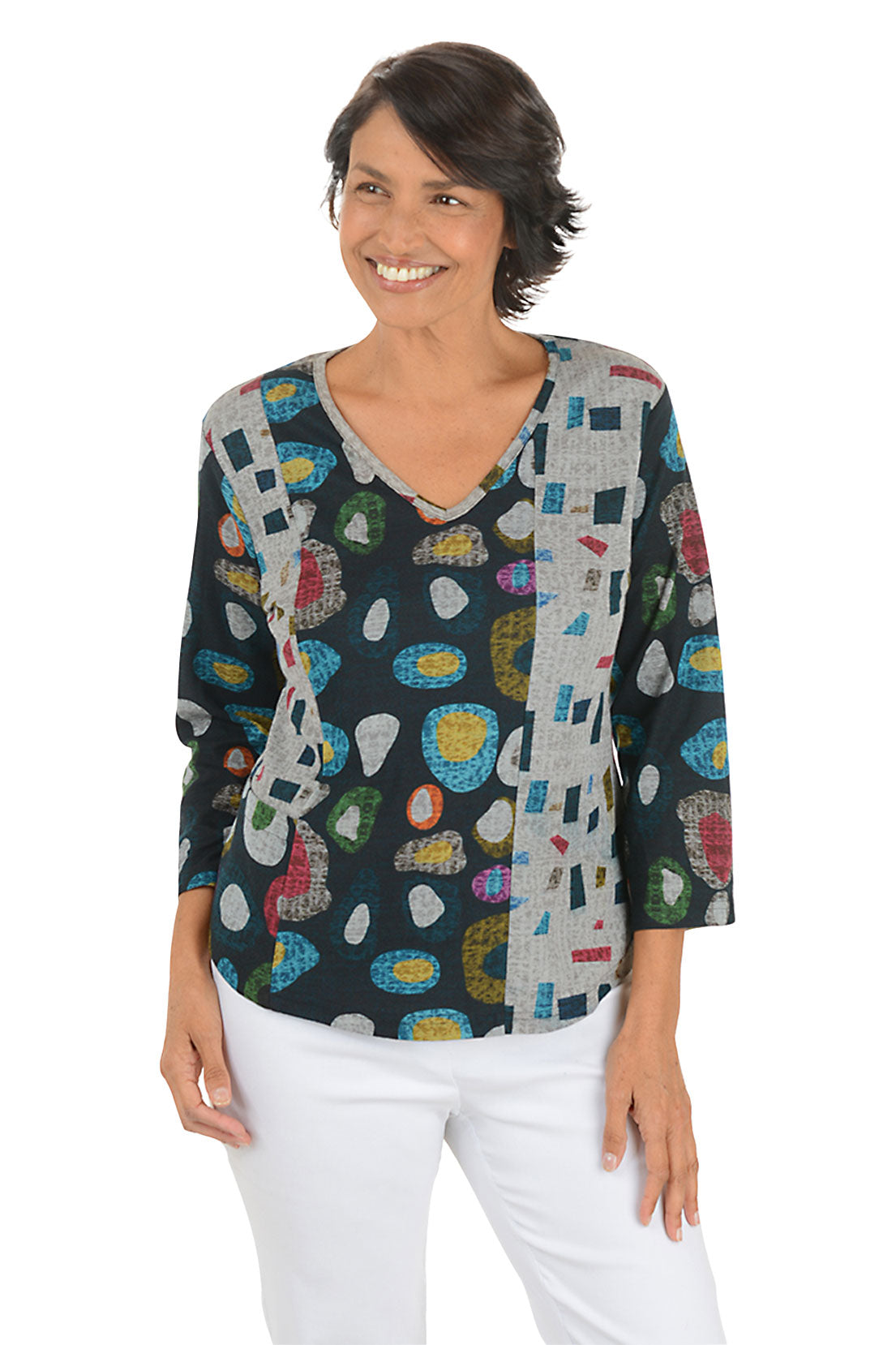 Woman wearing a 3/4 sleeve v-neck  abstract designed patterned top with jewel tones with white pants on a white background.