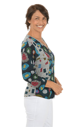 Woman wearing a 3/4 sleeve v-neck  abstract designed patterned top with jewel tones with white pants on a white background. Side shot.