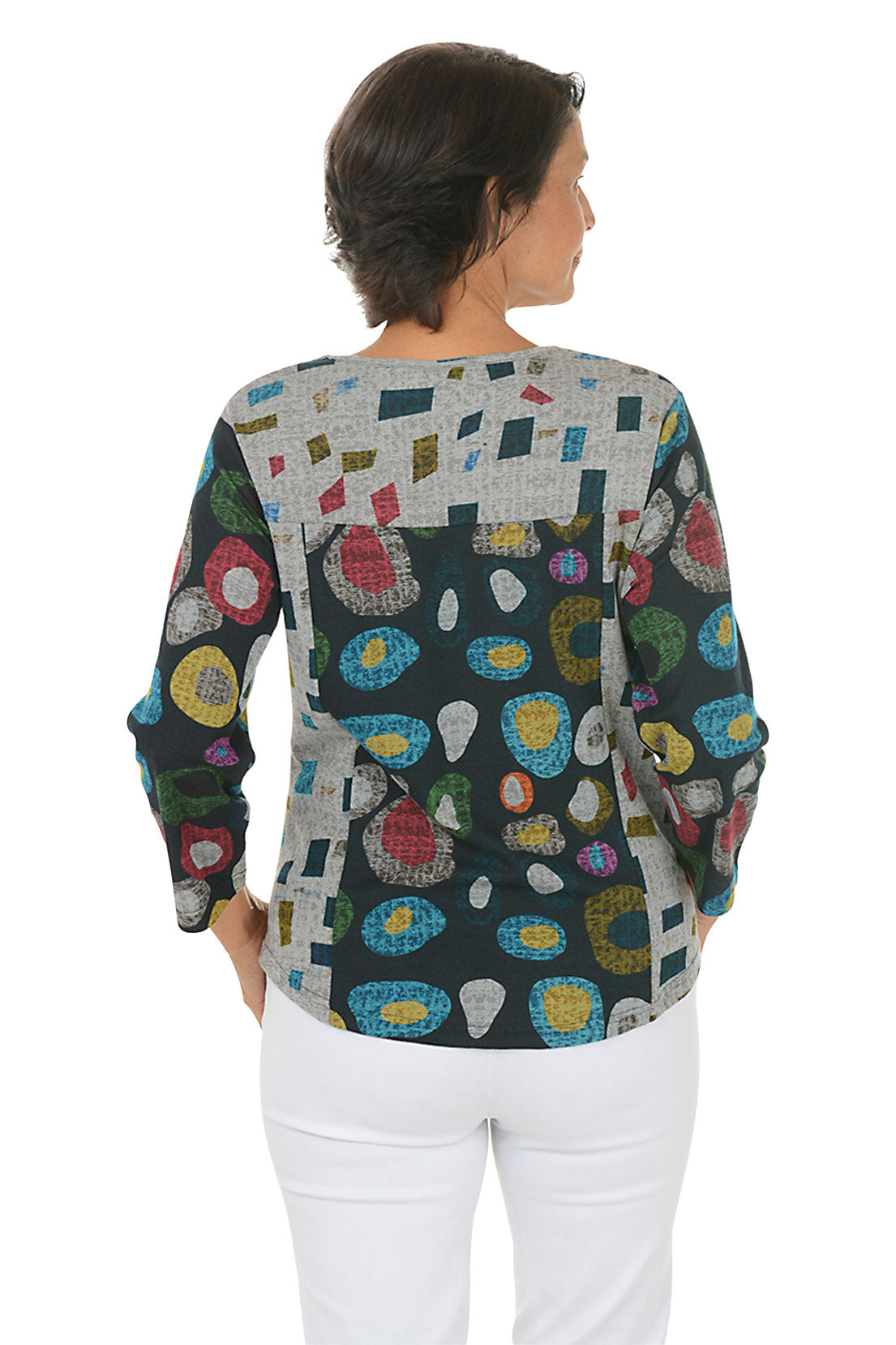 Woman wearing a 3/4 sleeve v-neck  abstract designed patterned top with jewel tones with white pants on a white background. Back shot.