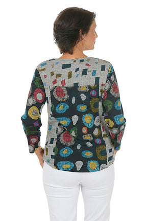 Woman wearing a 3/4 sleeve v-neck  abstract designed patterned top with jewel tones with white pants on a white background. Back shot.