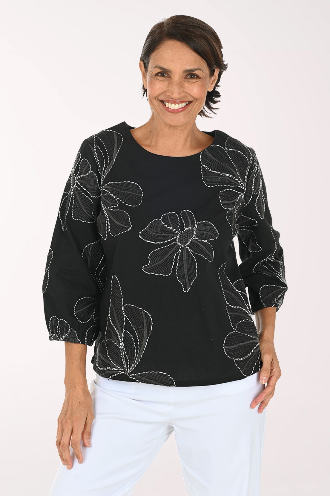 A smiling woman wearing a black three-quarter sleeve top with white floral embroidery, paired with white pants, against a plain white background.