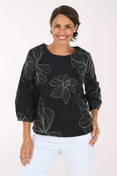 A smiling woman wearing a black three-quarter sleeve top with white floral embroidery, paired with white pants, against a plain white background.