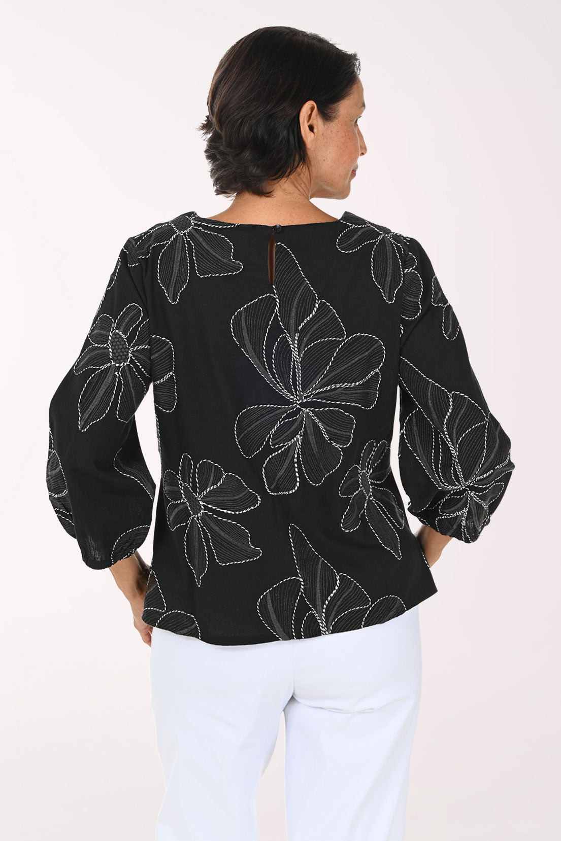 A smiling woman wearing a black three-quarter sleeve top with white floral embroidery, paired with white pants, against a plain white background.