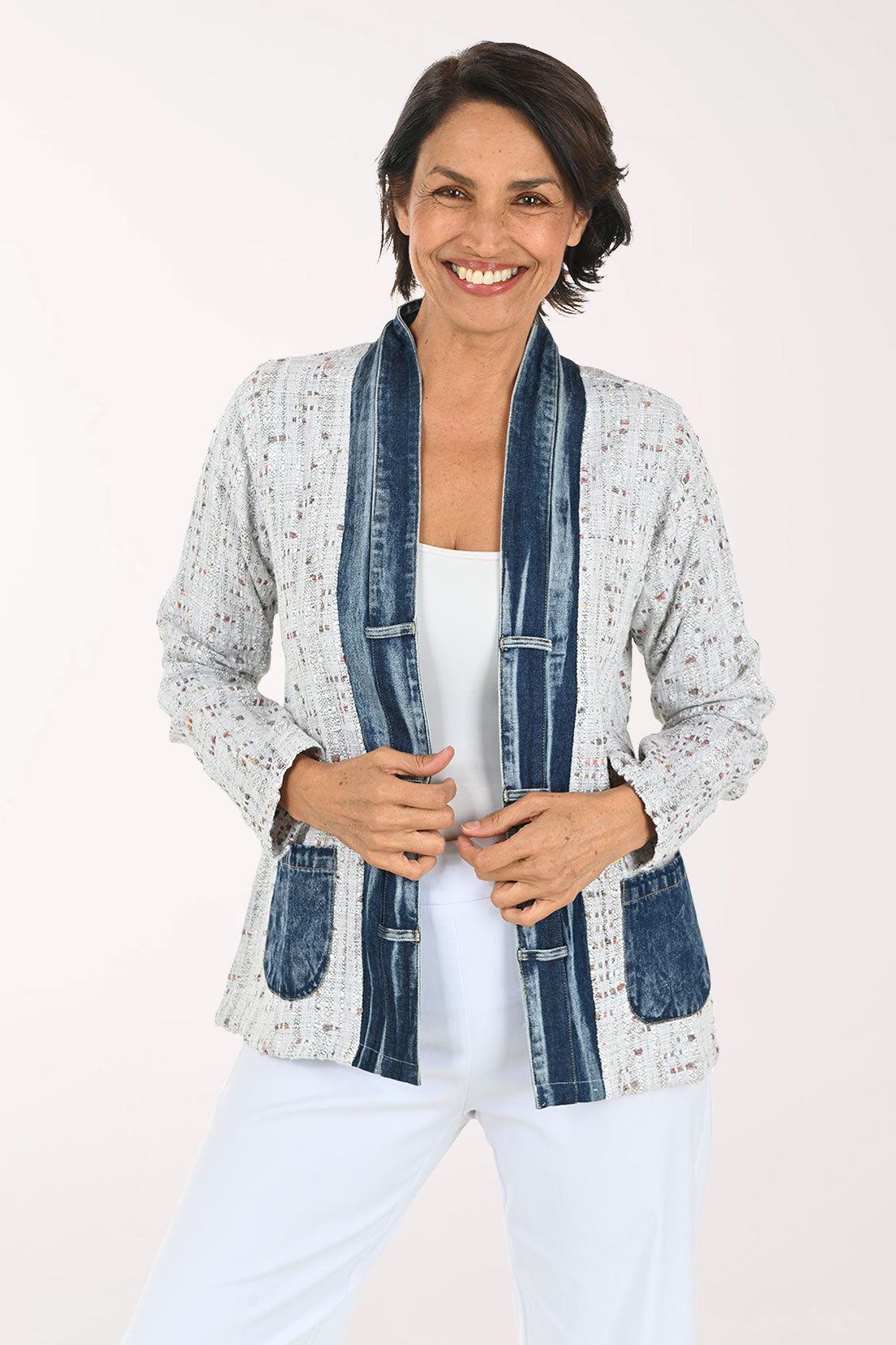 A smiling woman wearing a light gray speckled open-front cardigan with dark blue denim-style trim and front pockets, paired with a white top and white pants, against a plain light background.