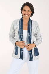 A smiling woman wearing a light gray speckled open-front cardigan with dark blue denim-style trim and front pockets, paired with a white top and white pants, against a plain light background.