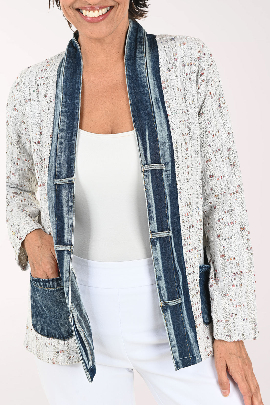 A smiling woman wearing a light gray speckled open-front cardigan with dark blue denim-style trim and front pockets, paired with a white top and white pants, against a plain light background.