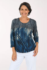A smiling woman wearing a blue and gray tie-dye long-sleeve top with white pants, standing against a plain white studio background.