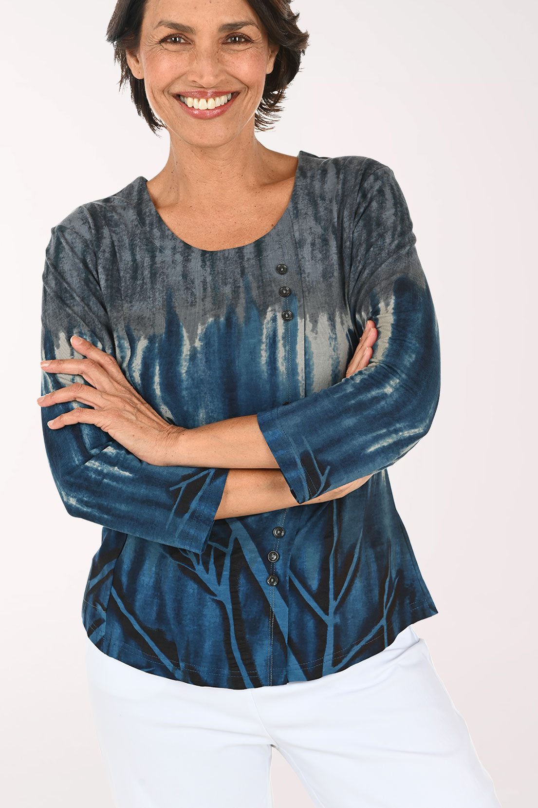 A smiling woman wearing a blue and gray tie-dye long-sleeve top with white pants, standing against a plain white studio background.
