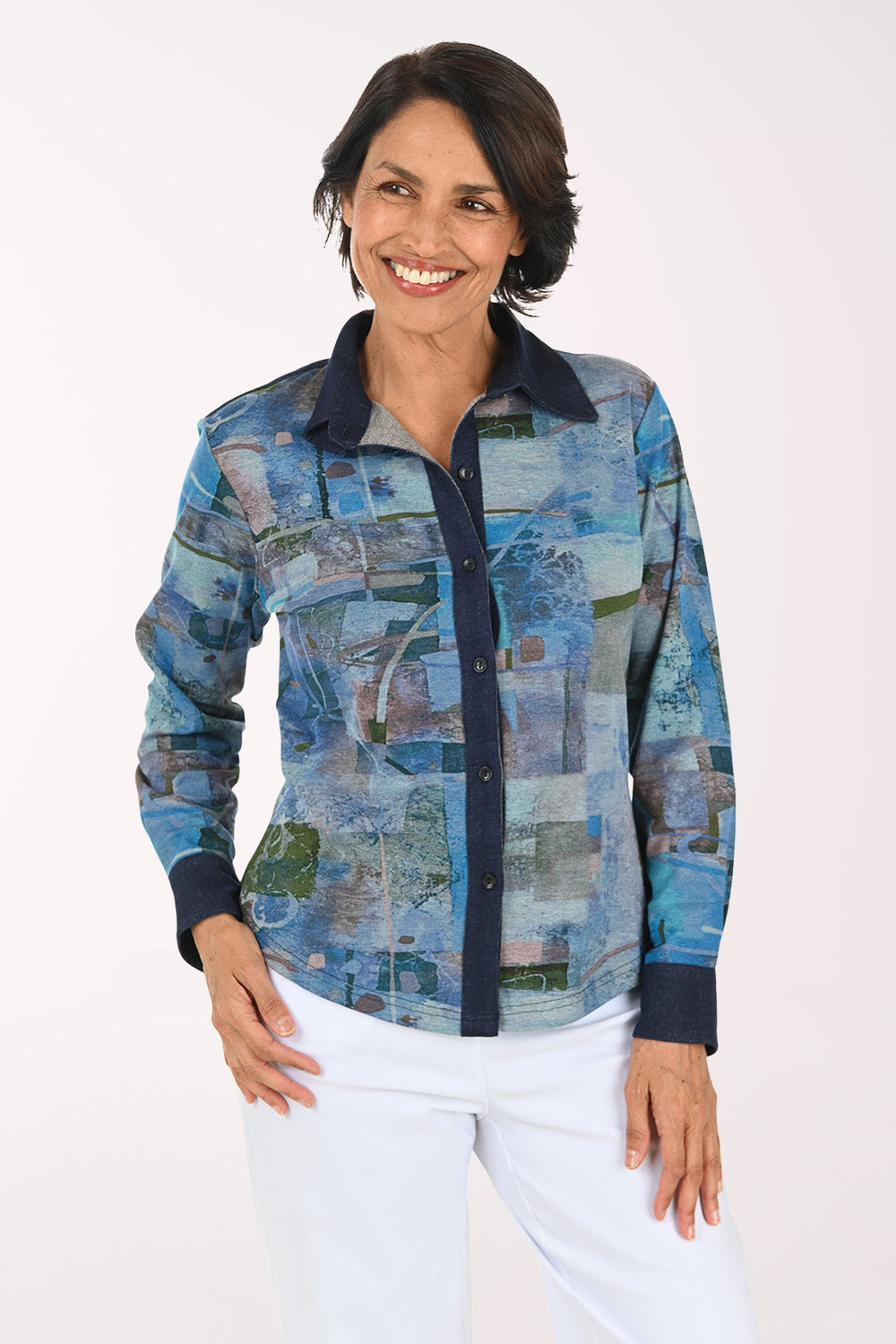 A smiling woman wearing a long-sleeve button-up shirt with an abstract blue, gray, and green patchwork print, paired with white pants, against a plain light background.