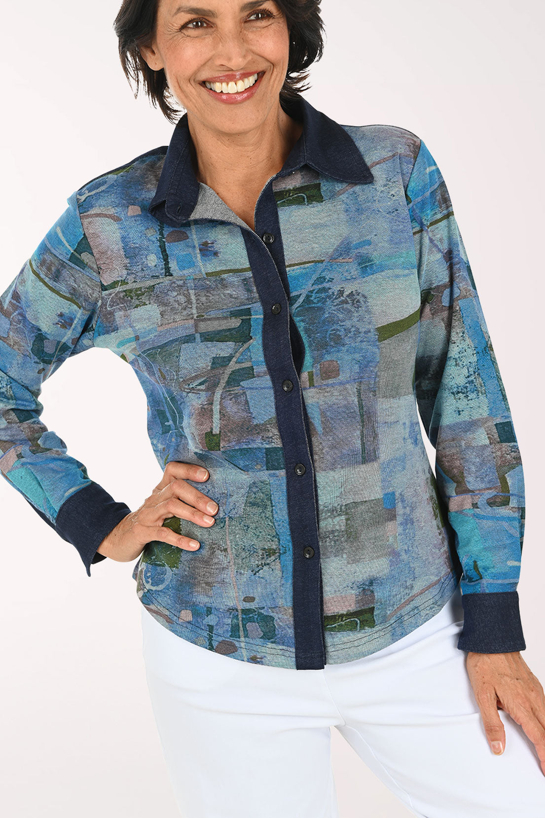 A smiling woman wearing a long-sleeve button-up shirt with an abstract blue, gray, and green patchwork print, paired with white pants, against a plain light background.