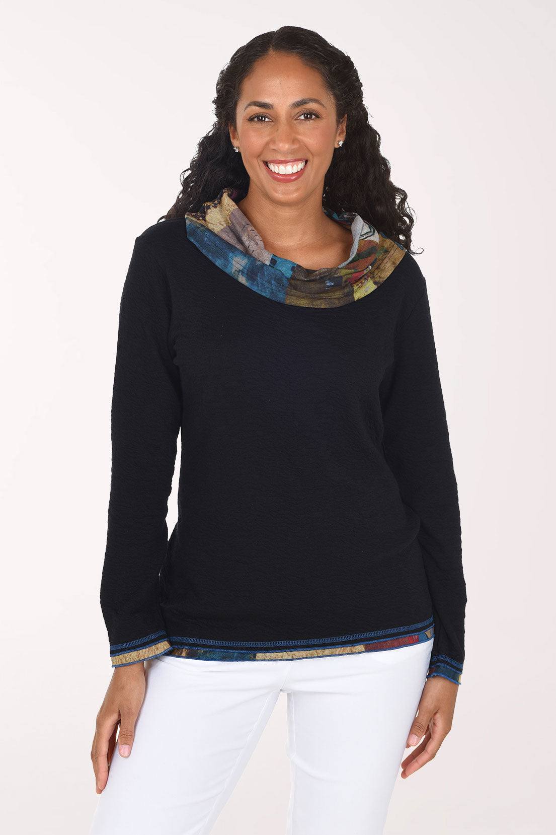 A smiling woman wearing a black long-sleeve sweater with a multicolor patterned cowl neck and hem trim, paired with white pants, against a plain white studio background.